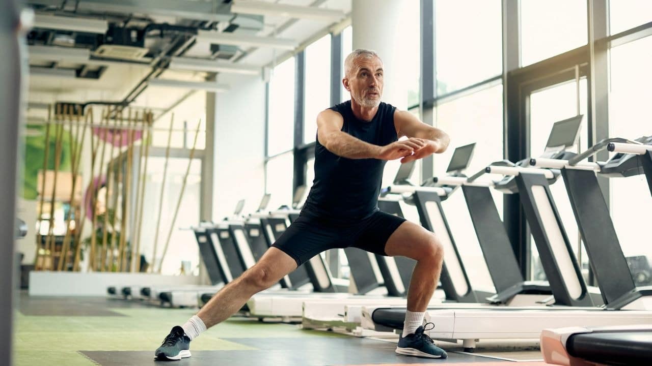 A fit, older man in activewear performs a lunge in a modern gym with treadmills.