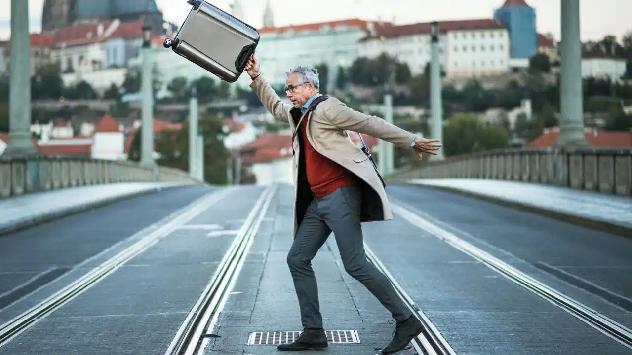 A man in a coat and glasses throws a suitcase in the air on a bridge.