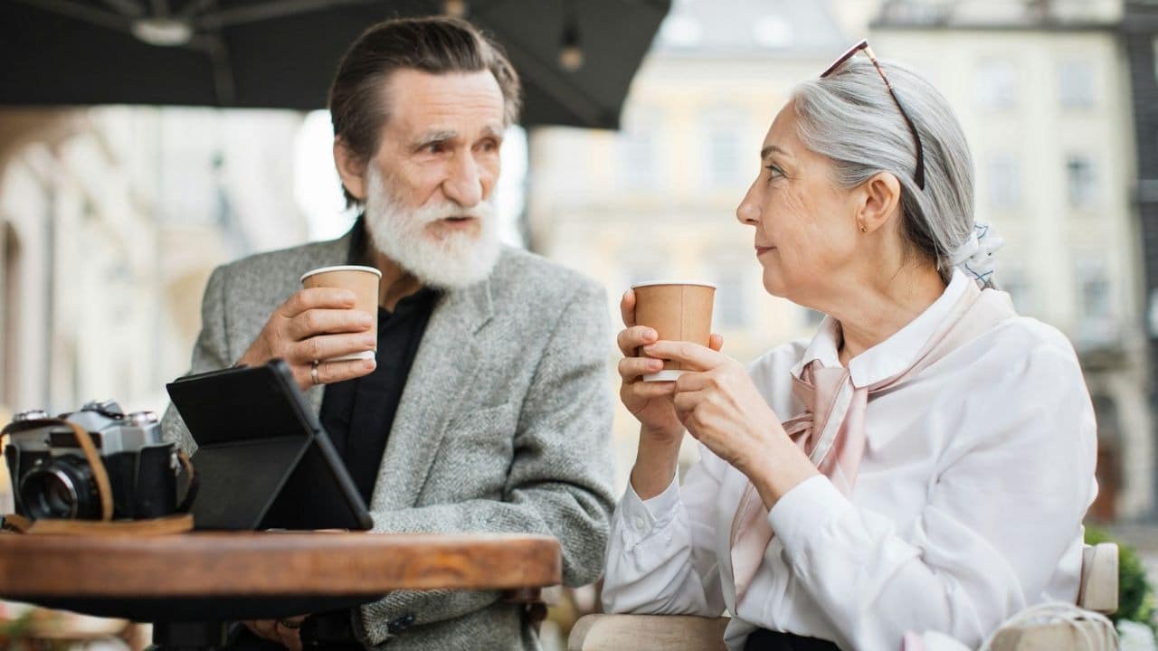 A senior couple sits at an outdoor cafe table, each holding a cup and looking at each other, with a camera and tablet nearby.