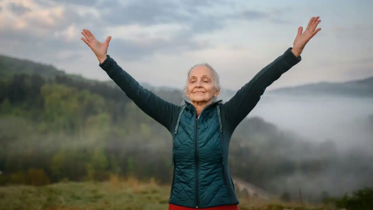 A smiling senior woman with outstretched arms embraces nature in a misty landscape.