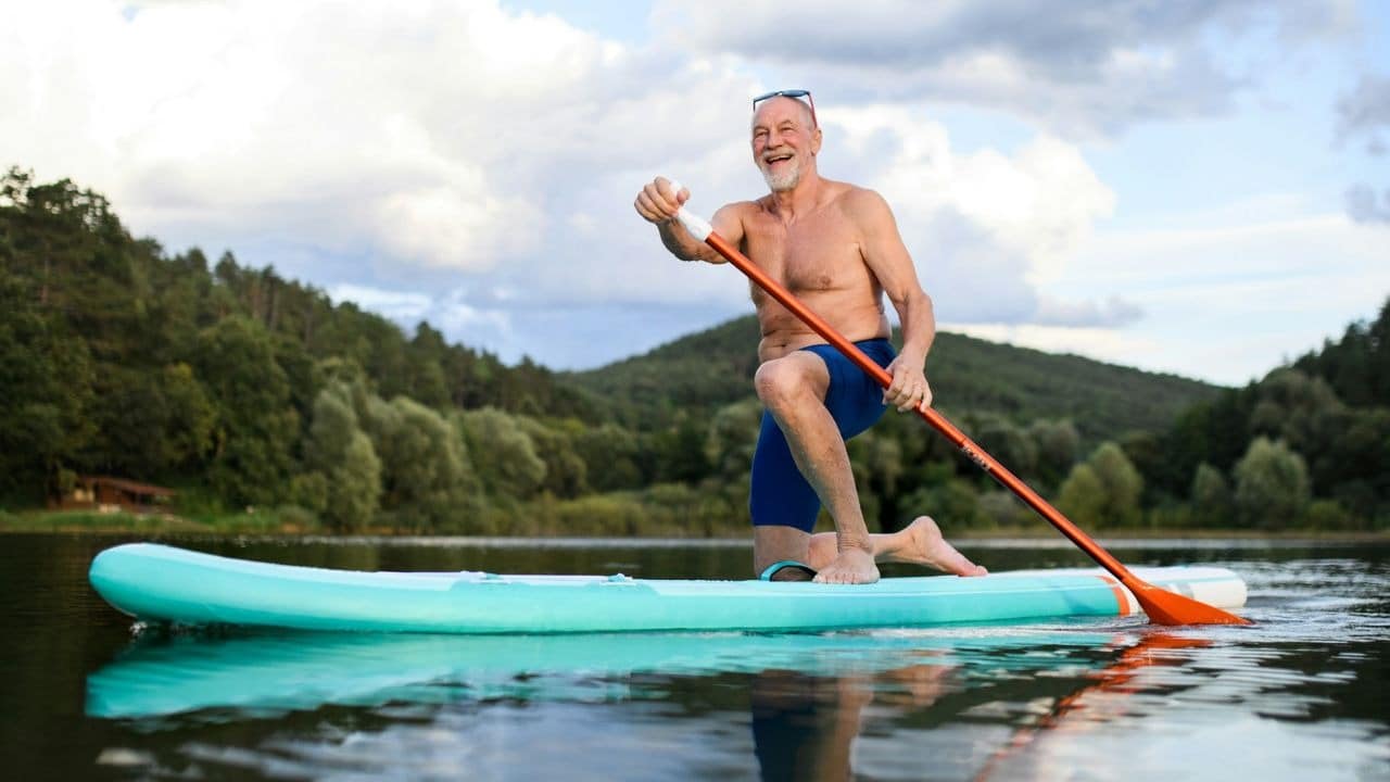 A shirtless, smiling older man on a paddleboard on a lake.