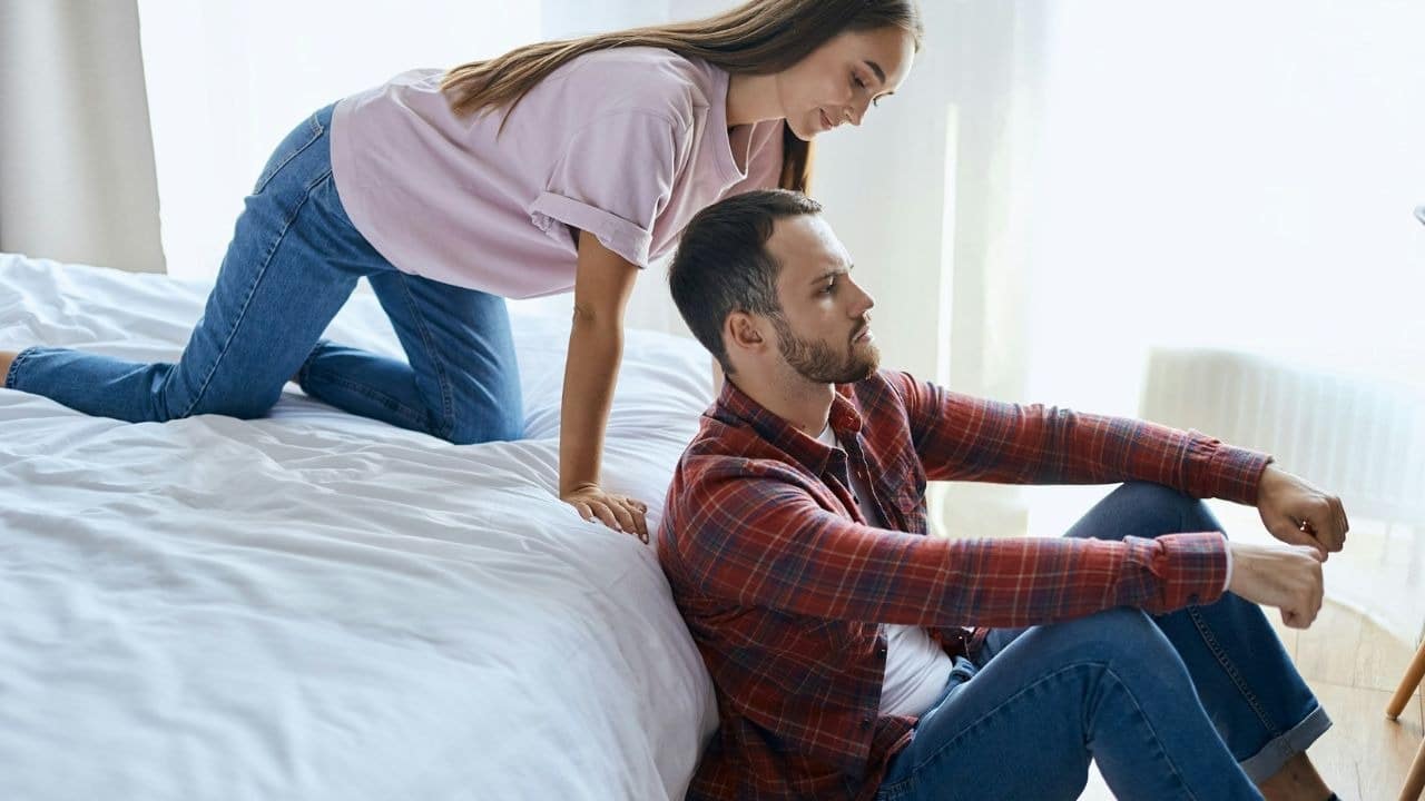 A man sits on the floor with his back to a woman kneeling on a bed.