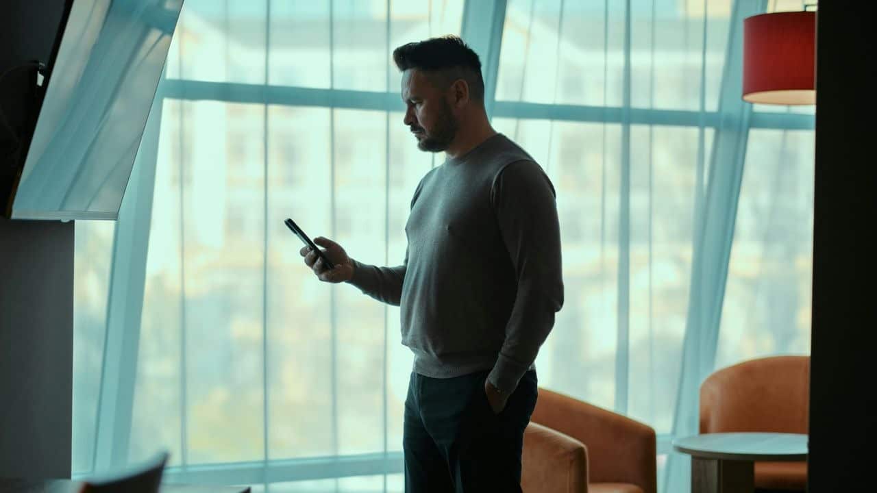 A man stands silhouetted against a large window, looking at his phone.