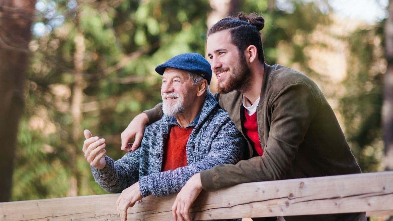 A cheerful older man and younger man stand together outdoors, looking off into the distance.