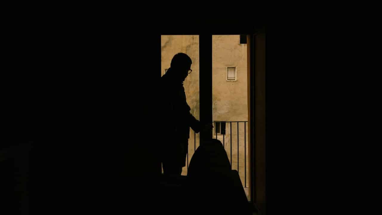A silhouetted person stands on a balcony, looking out at a building.