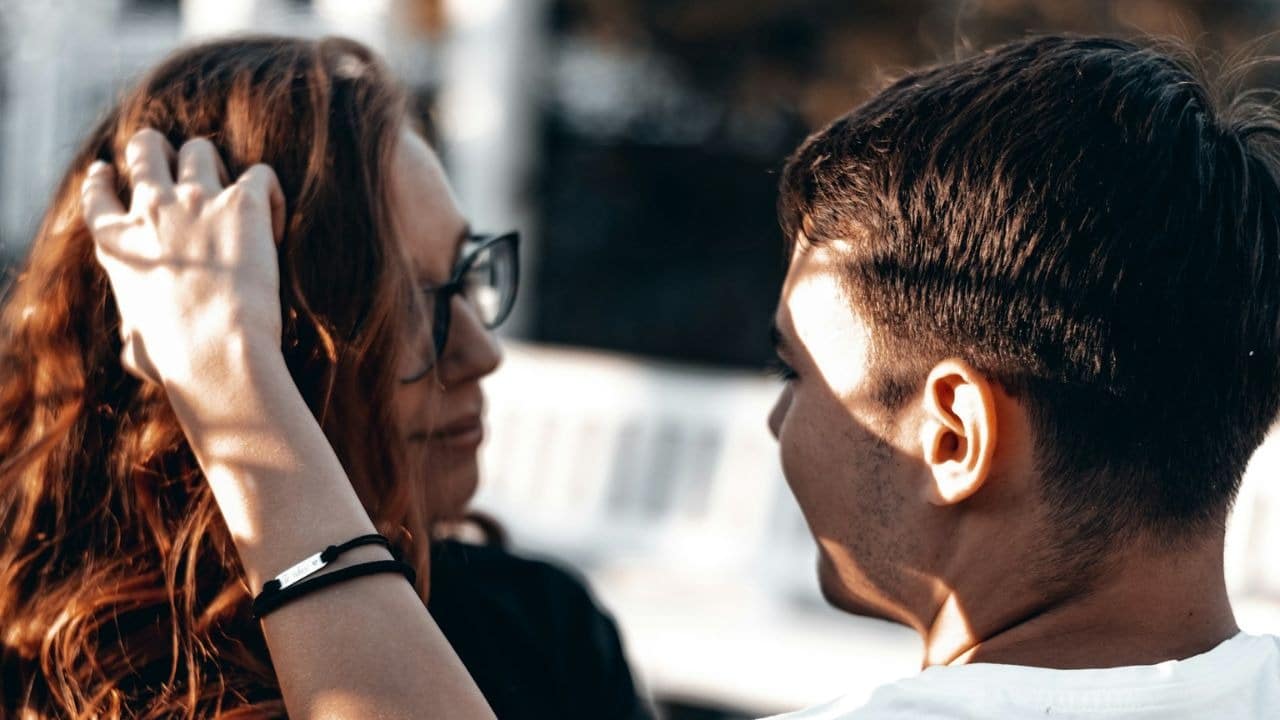 A woman with long, wavy hair and glasses looks at a man, with her hand near her ear.