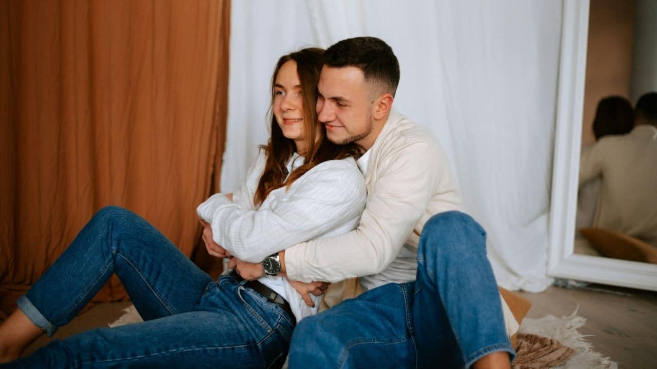 A man in a cream shirt and jeans hugs a woman in a white shirt and jeans from behind.