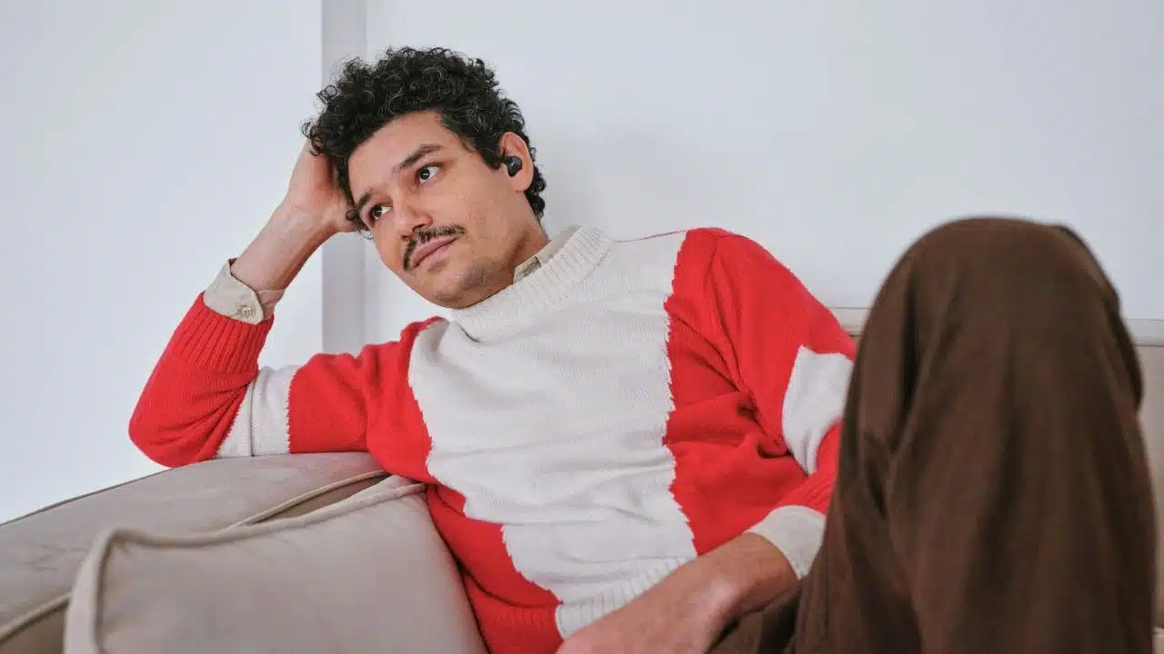 A man with curly hair and a mustache reclines on a couch, wearing a red and white sweater.