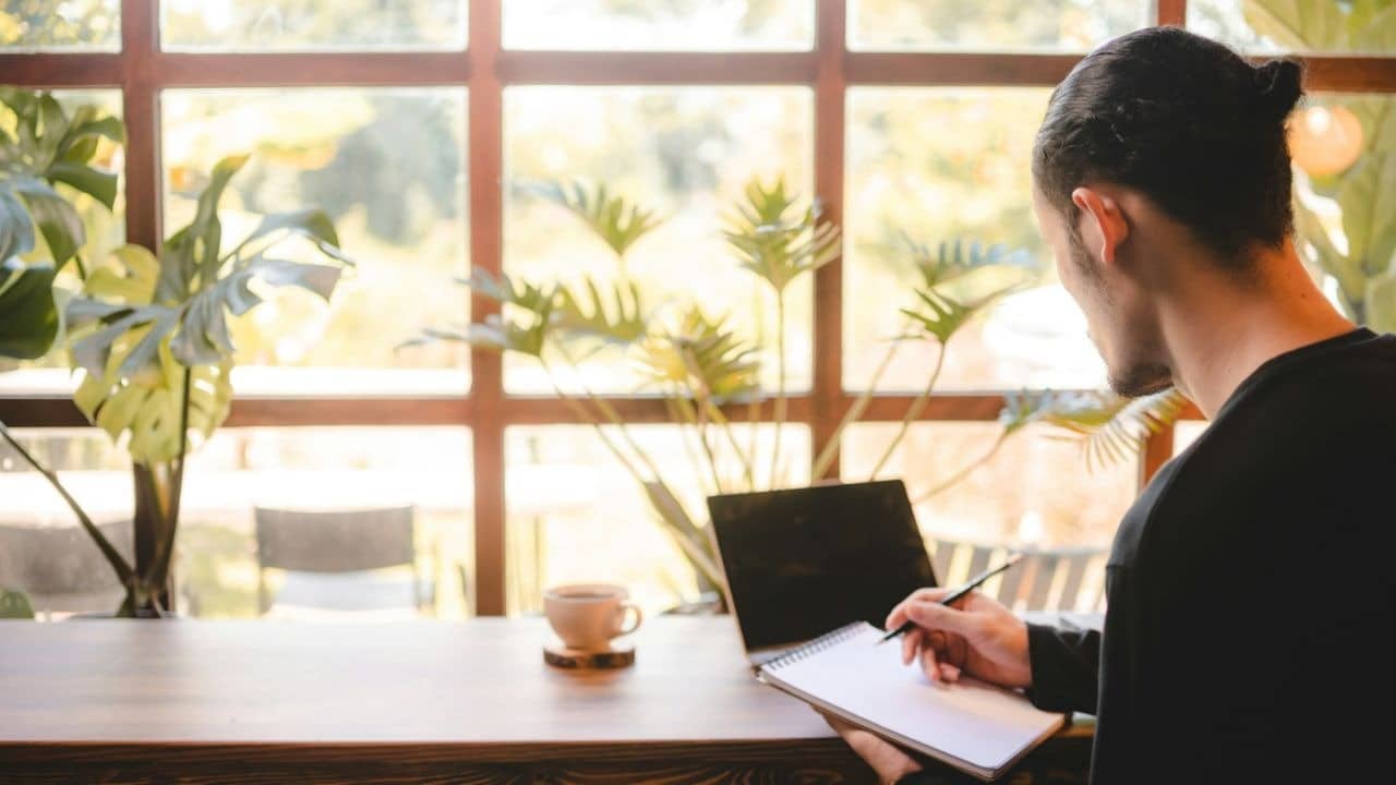 A man with his hair in a bun writes in a notebook at a desk with a laptop and coffee.