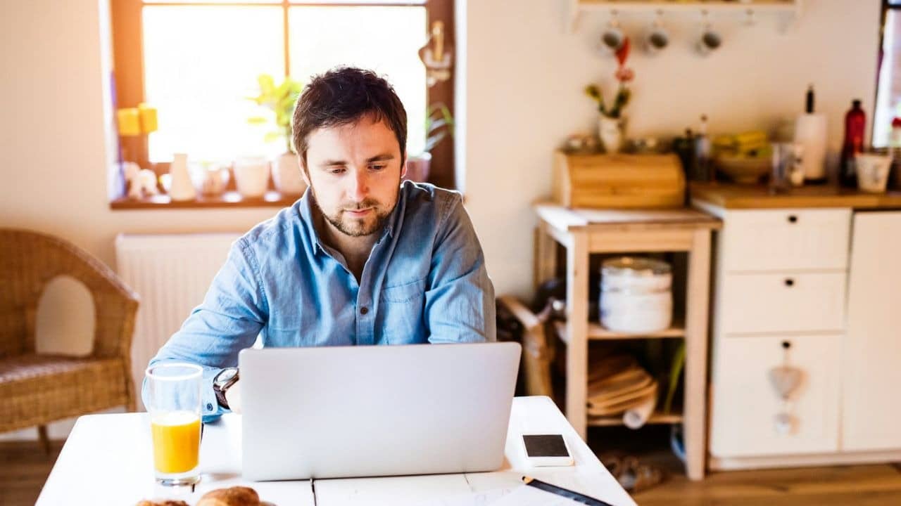 A man with a beard sits at a table, looking at a laptop, with a glass of juice.