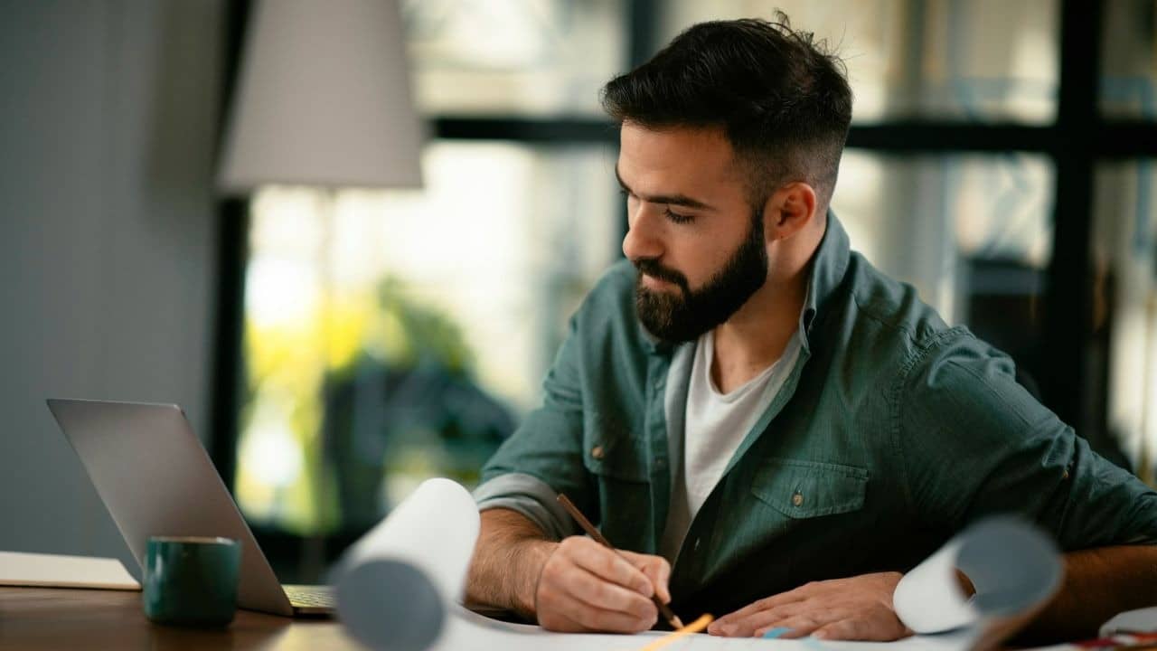 A bearded man in a green shirt draws or writes at a desk with a laptop and rolled papers.