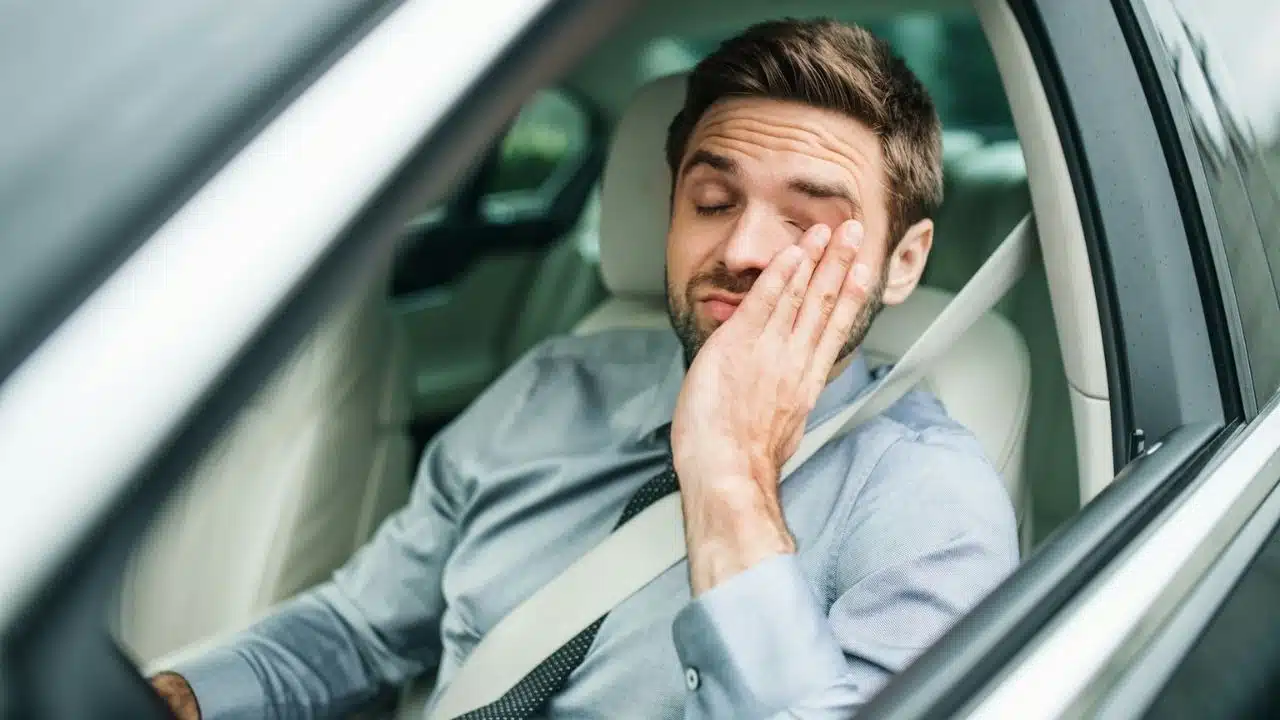 A man in a dress shirt and tie sits in a car, rubbing his eye with a tired expression.