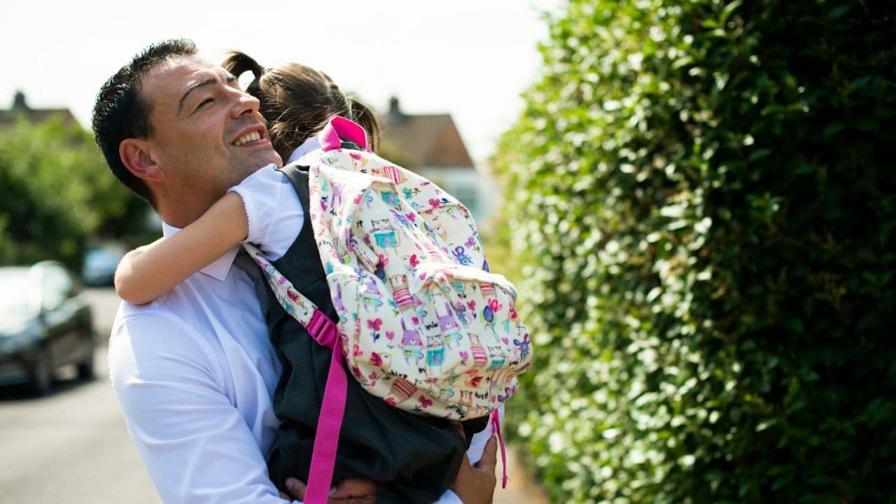 A man in a white shirt smiles as he hugs a young child with a backpack.