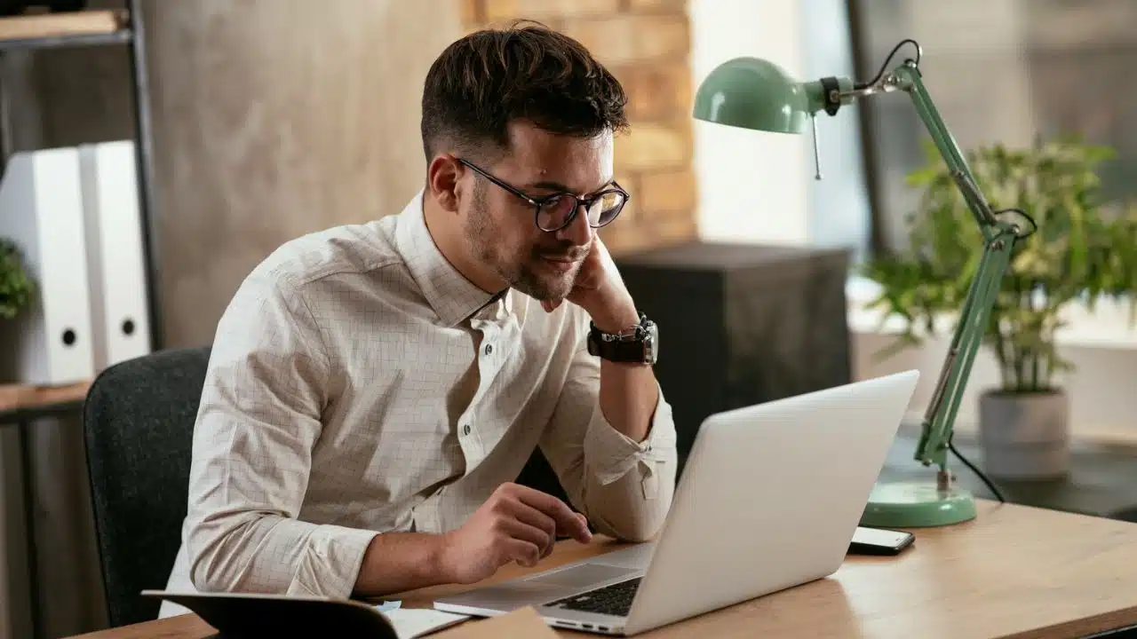 A man wearing glasses and a plaid shirt works at a laptop, with a green lamp on his desk.