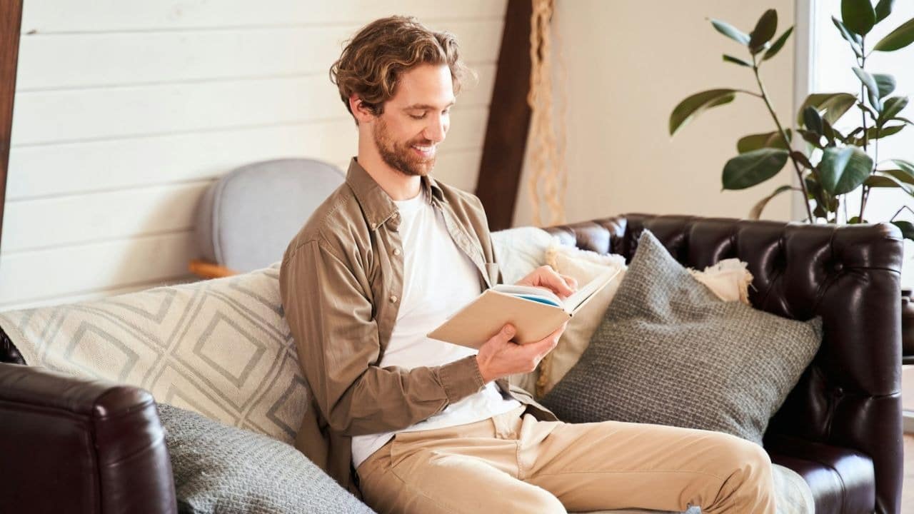 A man with wavy hair smiles while reading a book on a couch.