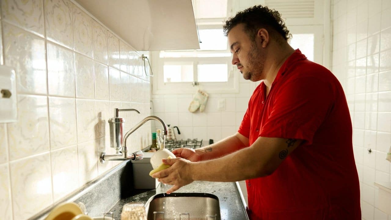A man in a red shirt washes dishes at a kitchen sink.