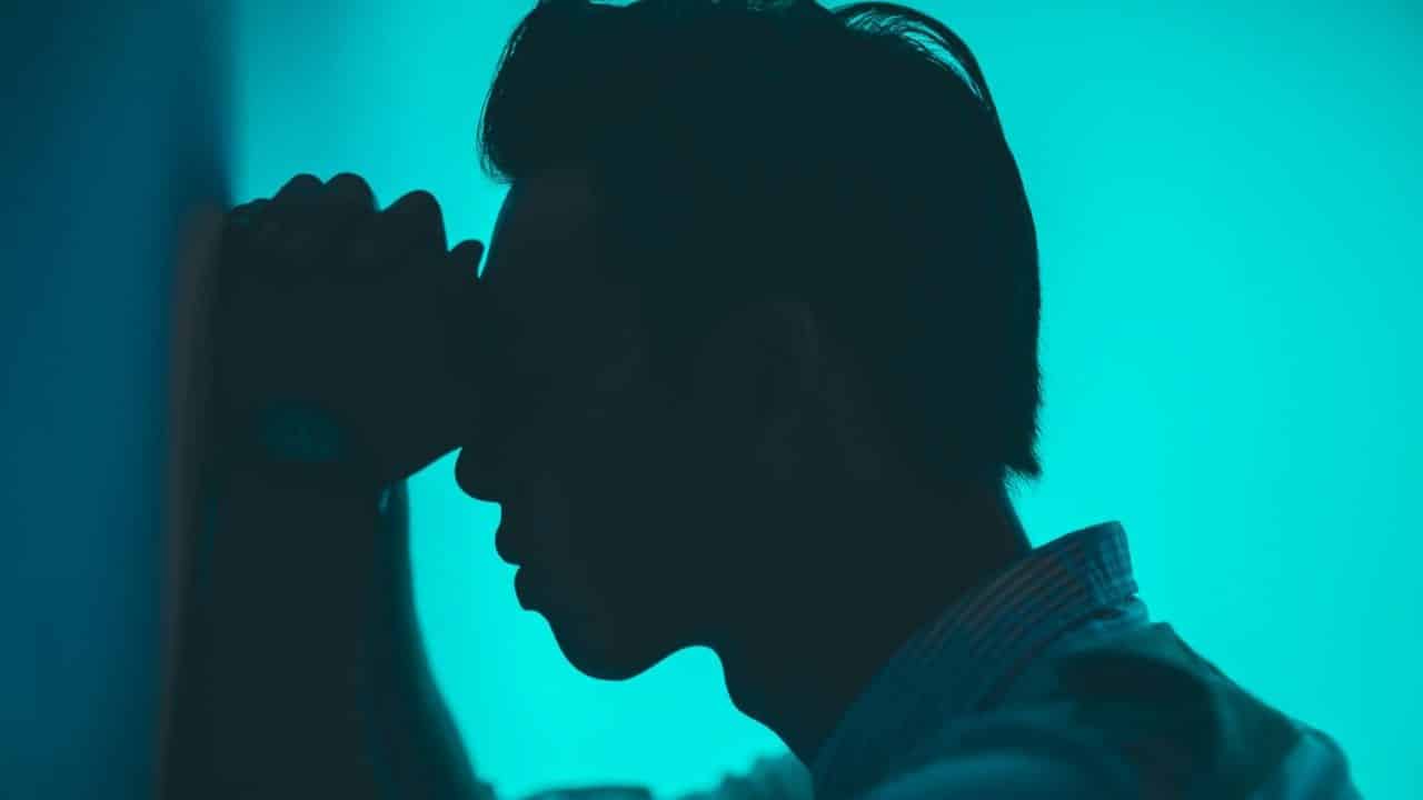 A person in silhouette holds their head in their hands against a blue background.