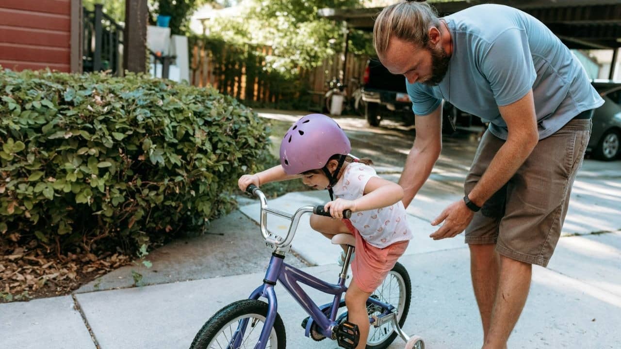 A man helps a young child in a purple helmet learn to ride a bicycle.