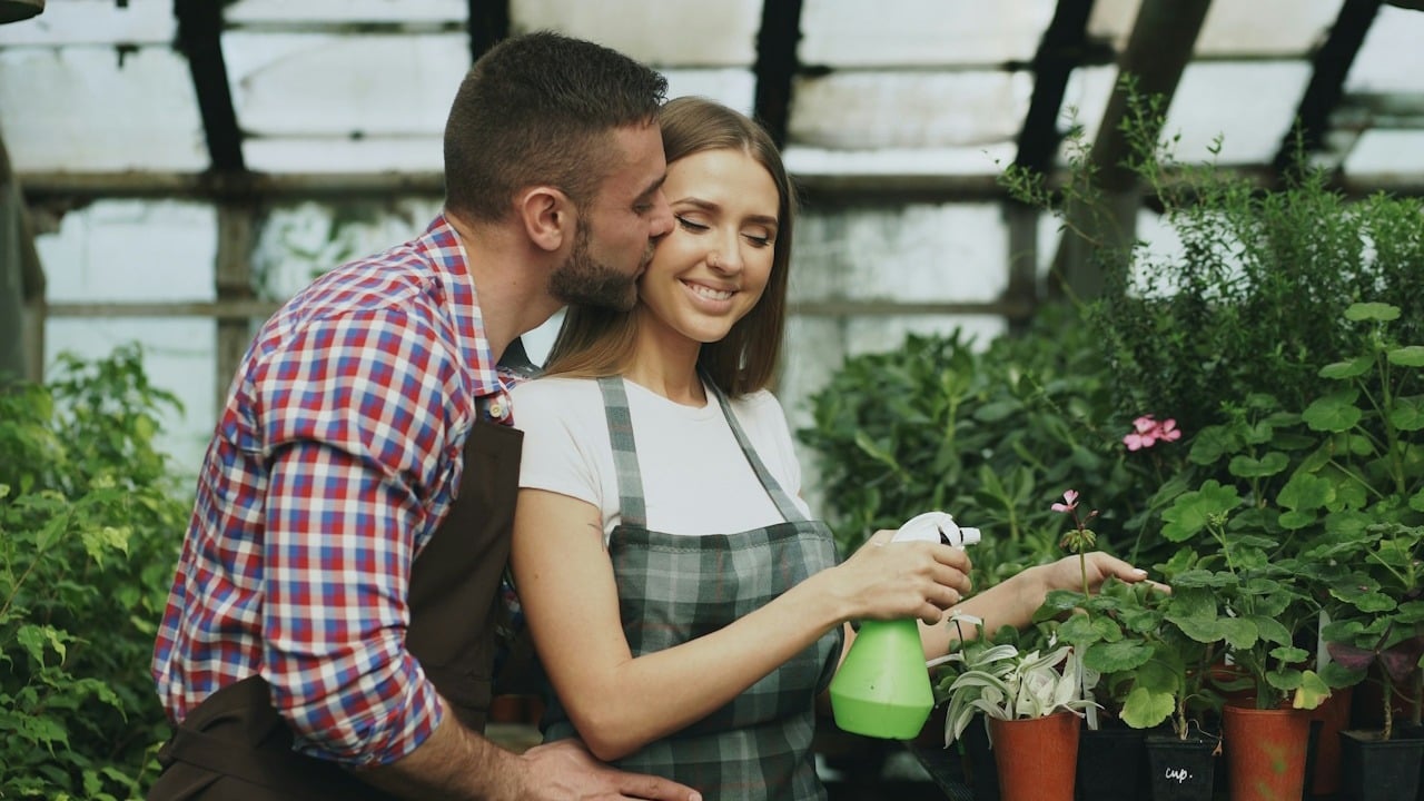 A man kissing a woman who’s tending to some plants in a greenhouse.