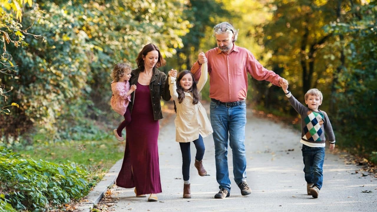 A family of four, two adults and two children, walk hand-in-hand on a path surrounded by trees.