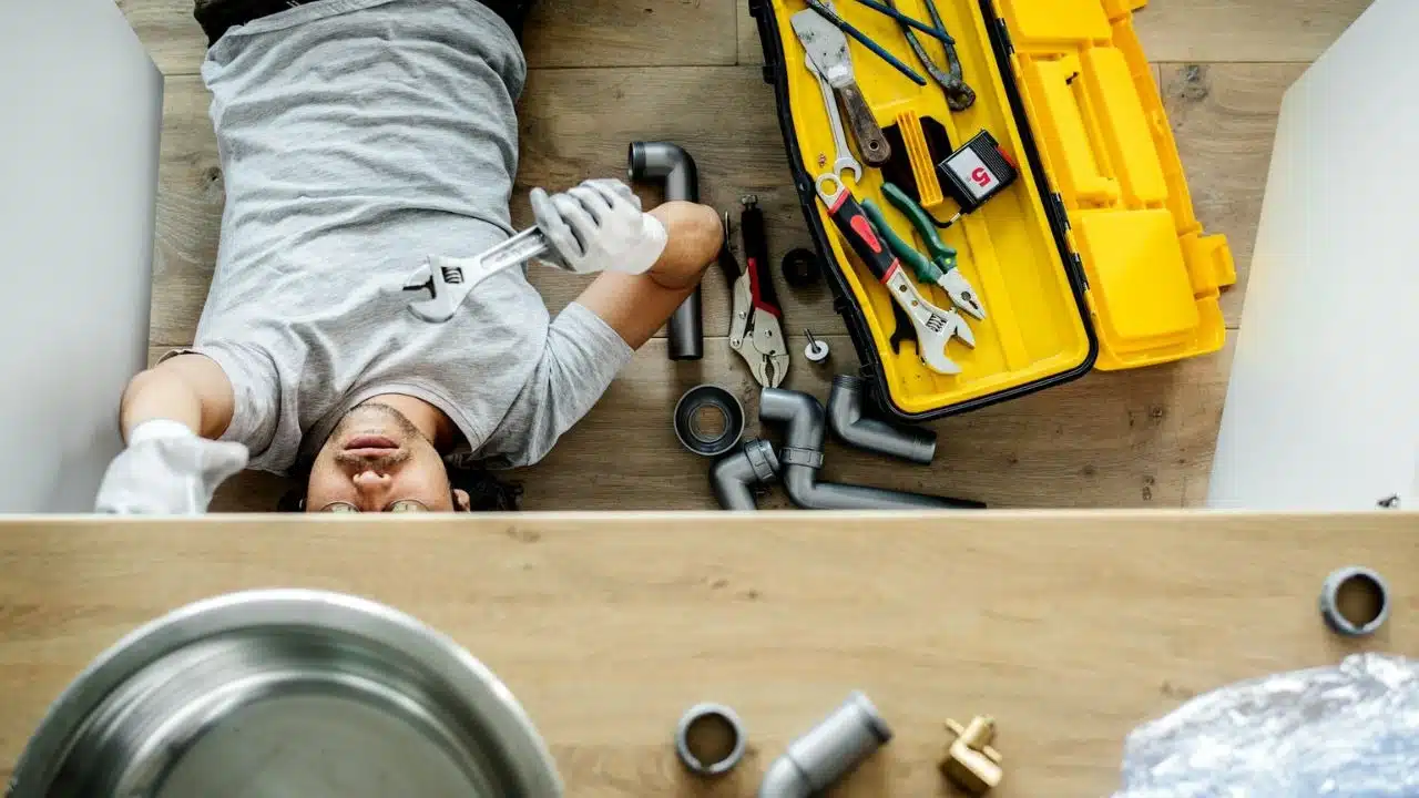 A man lies on his back under a sink, holding a wrench, with a toolbox nearby.