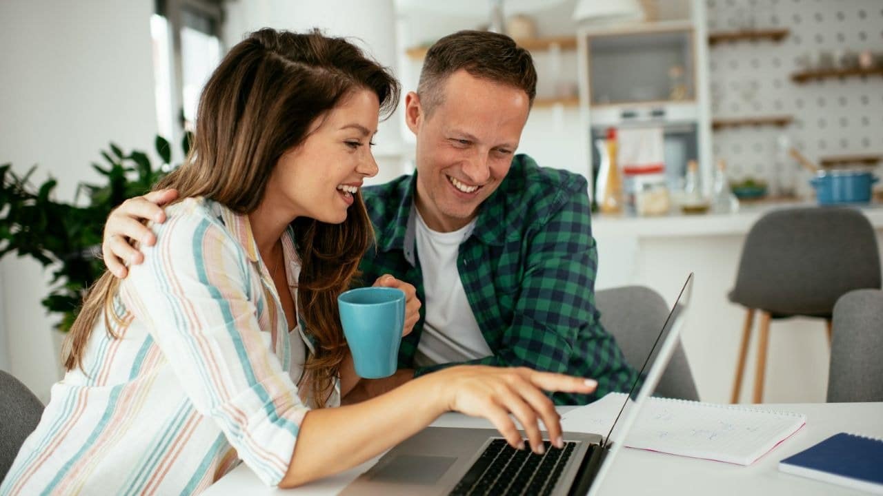 A man and a woman smile while looking at a laptop together, with the woman pointing at the screen.