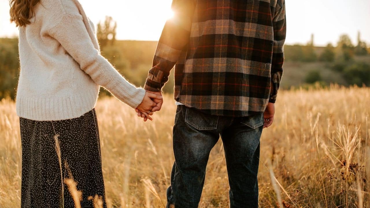 A man and woman hold hands in a field of tall grass during sunset.