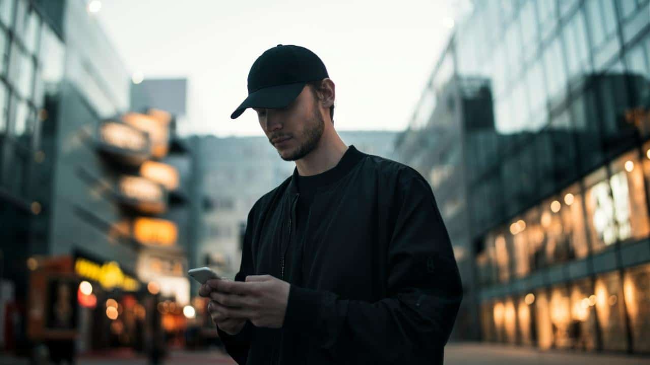 A man in a baseball cap checking his phone on a city street.