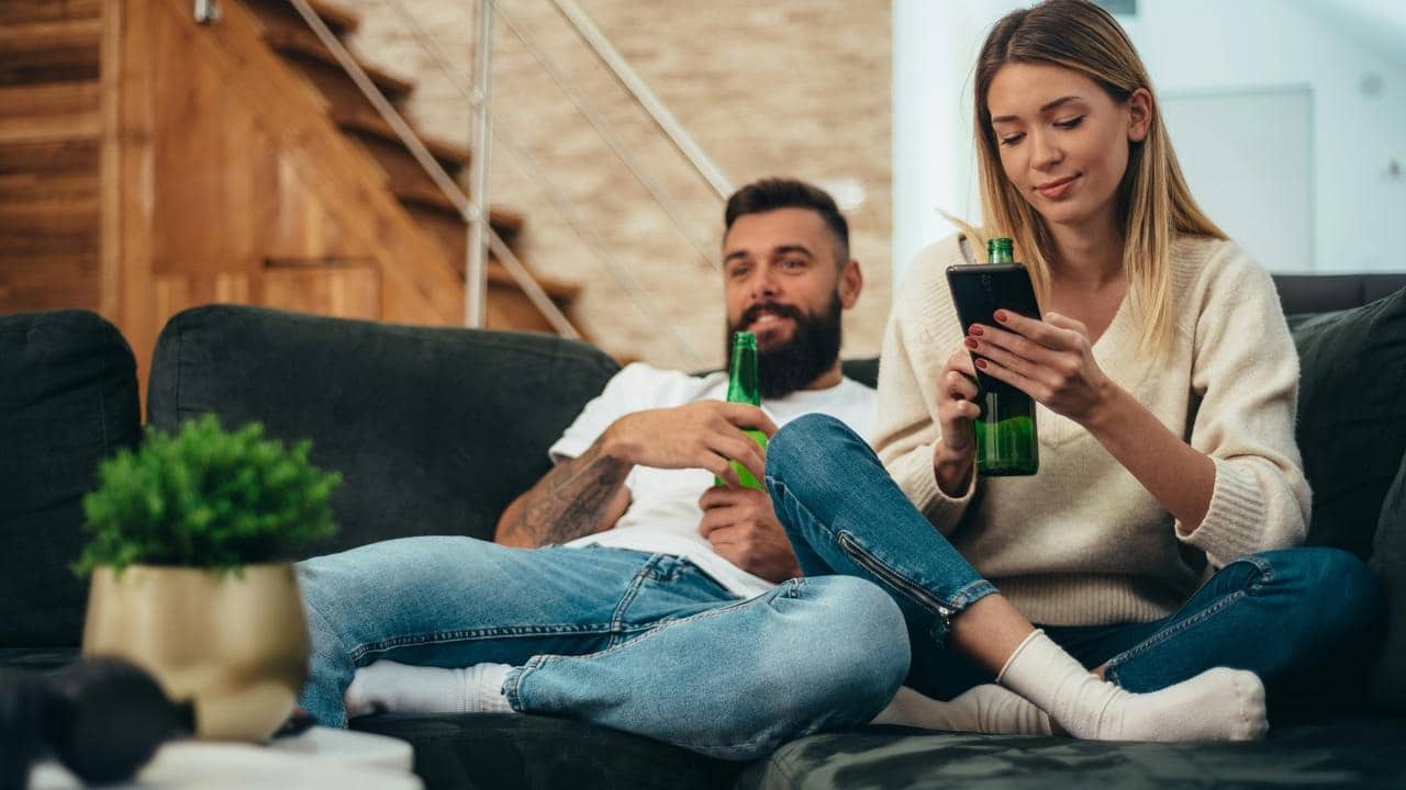 A couple relaxing on a couch with beer bottles as the woman checks her phone.