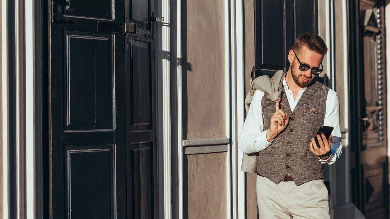 A man in sunglasses with a jacket over his shoulder checking his phone.