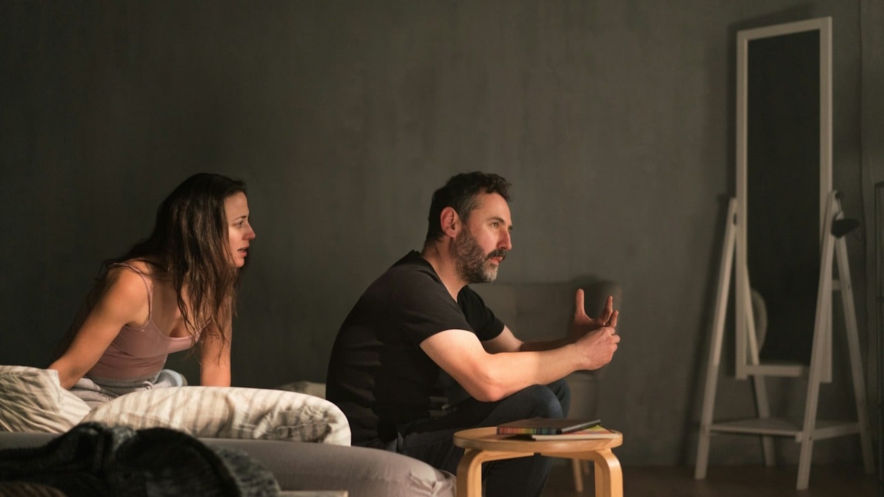 A woman talking to a man who has his back turned to her and refuses to answer as they sit on a bed.