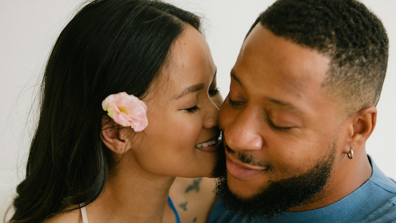 A woman with a flower in her ear about to kiss a bearded man’s cheek.