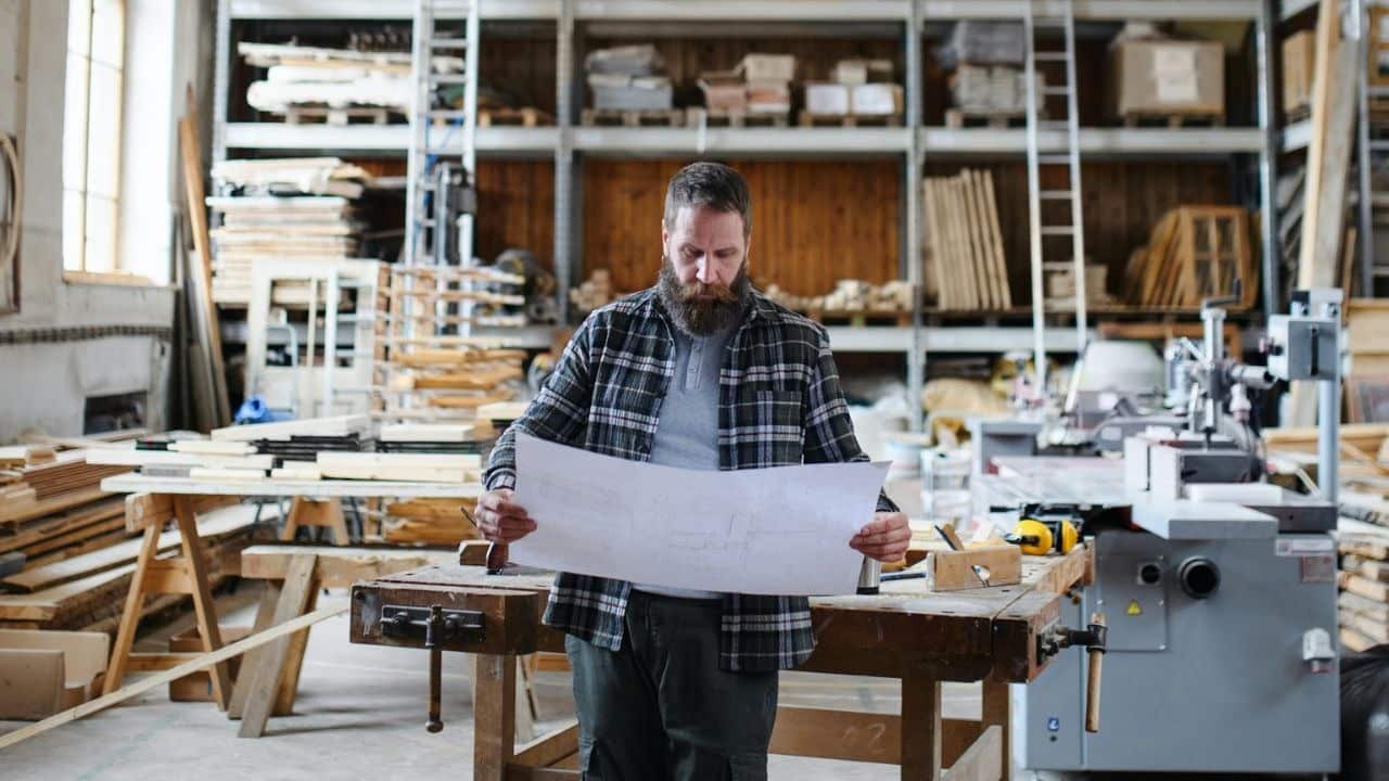 A bearded man in a plaid shirt examines blueprints in a woodworking shop.