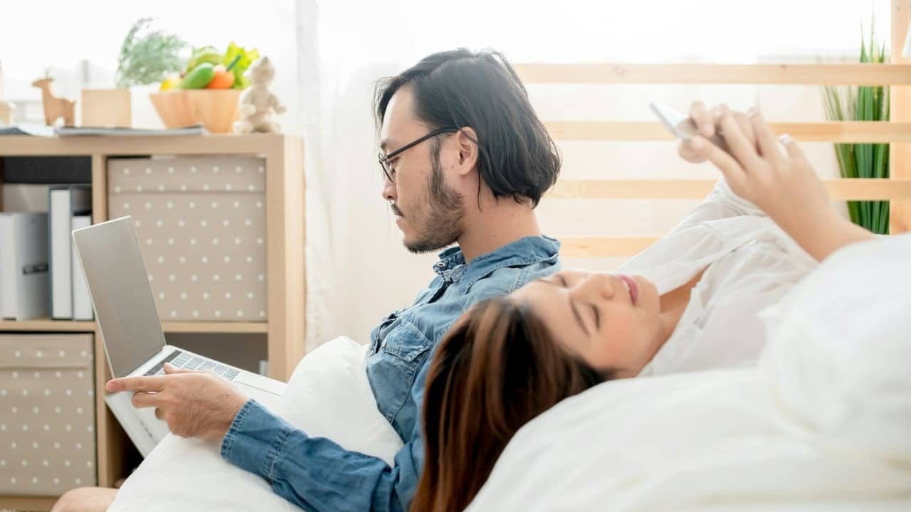 A man works on a laptop while a woman relaxes beside him in bed.