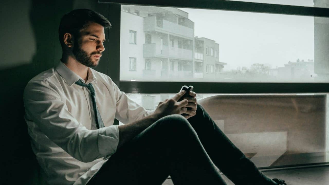 A pensive man sits by a window, looking at his phone.