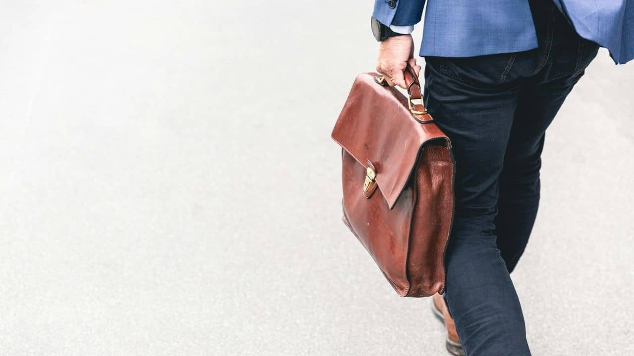 A man in a suit jacket and jeans walks carrying a brown briefcase.