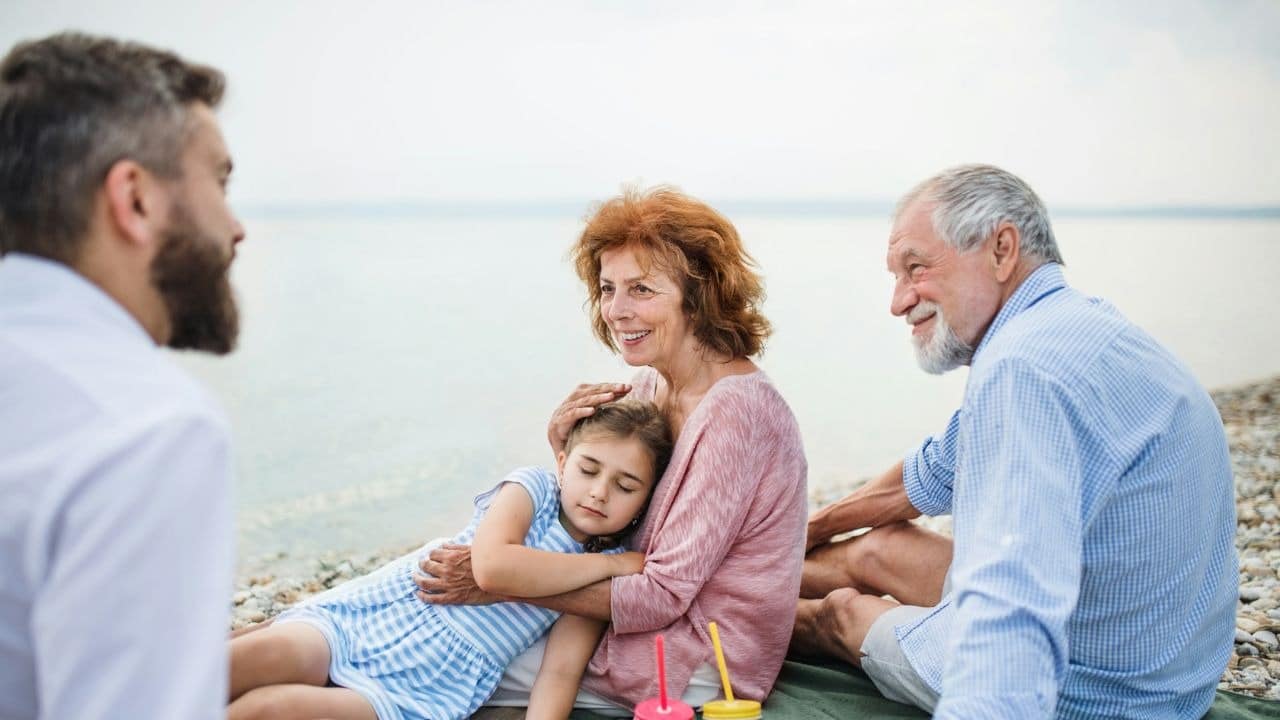 A grandmother embraces her granddaughter while a family sits by the water.