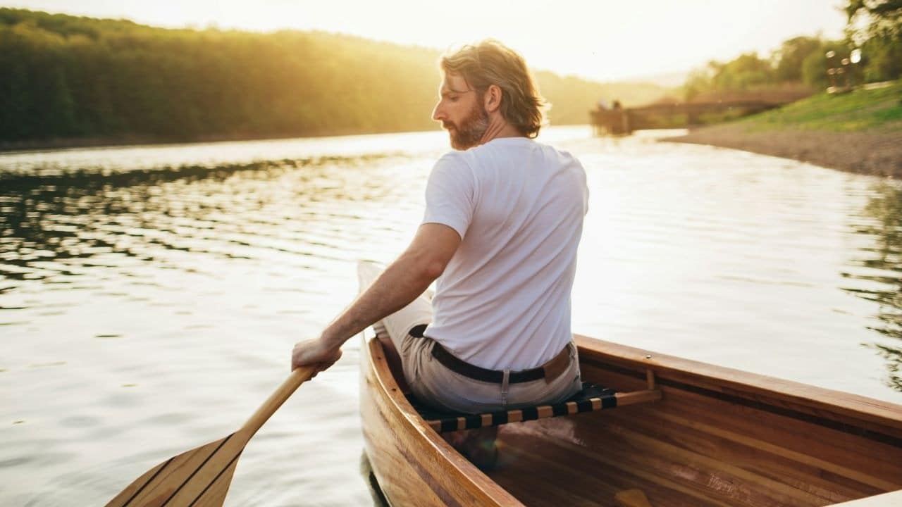 A man with a beard paddles a wooden canoe on a sunny lake.