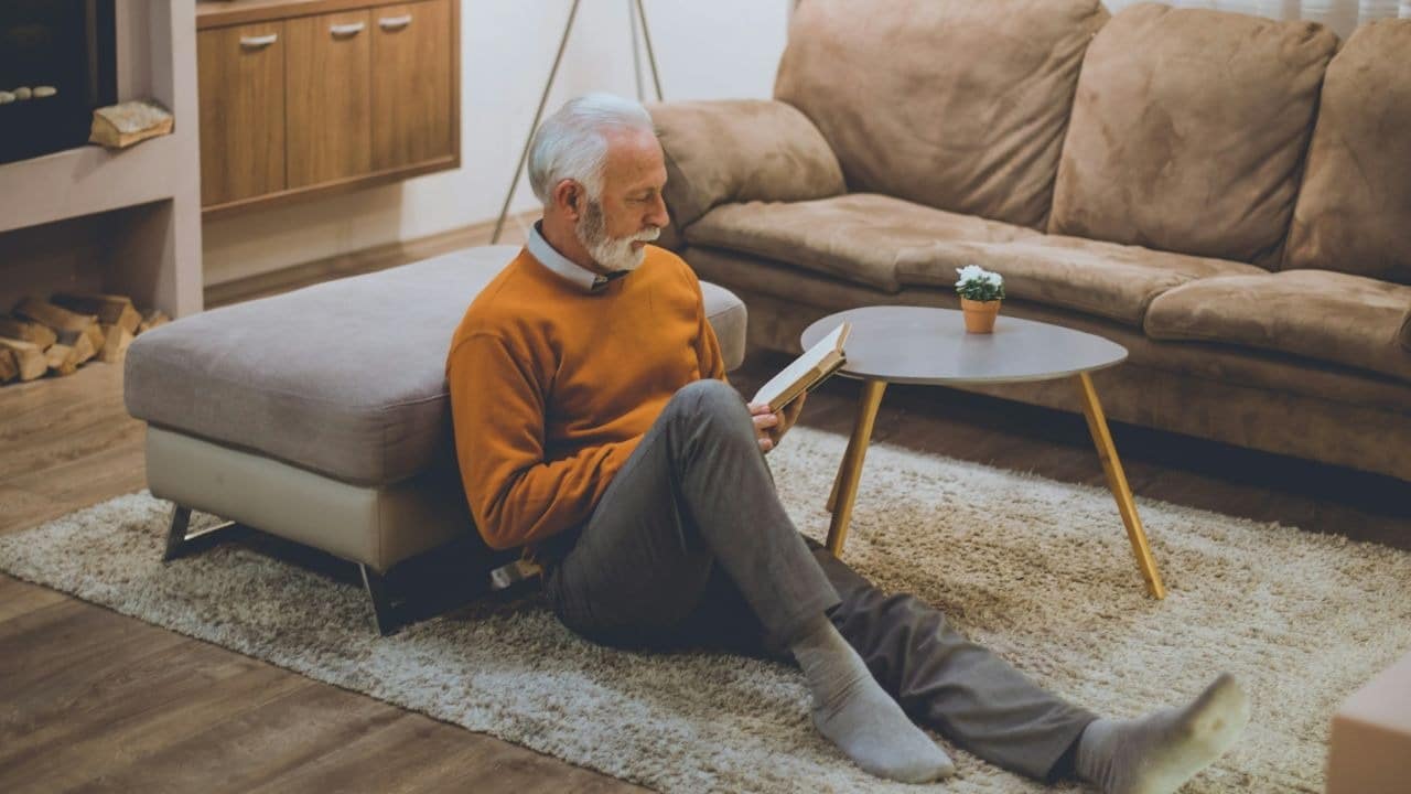 A gray-haired man in an orange sweater sits on the floor, reading.