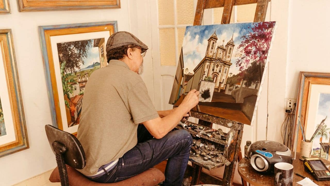 A male artist with a cap paints a building on a canvas in a room.