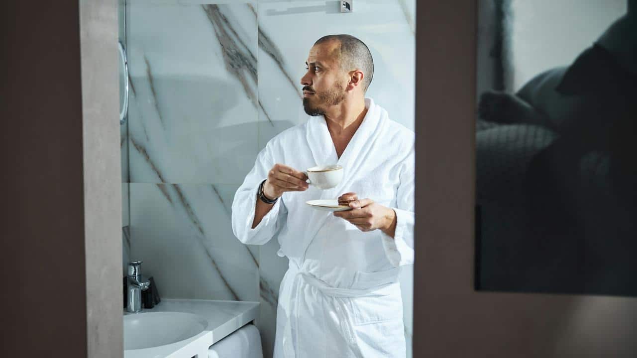 A man in a bathrobe holding a cup and saucer in a bathroom.