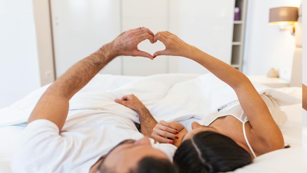 A couple lying on bed forming heart shape with hand.