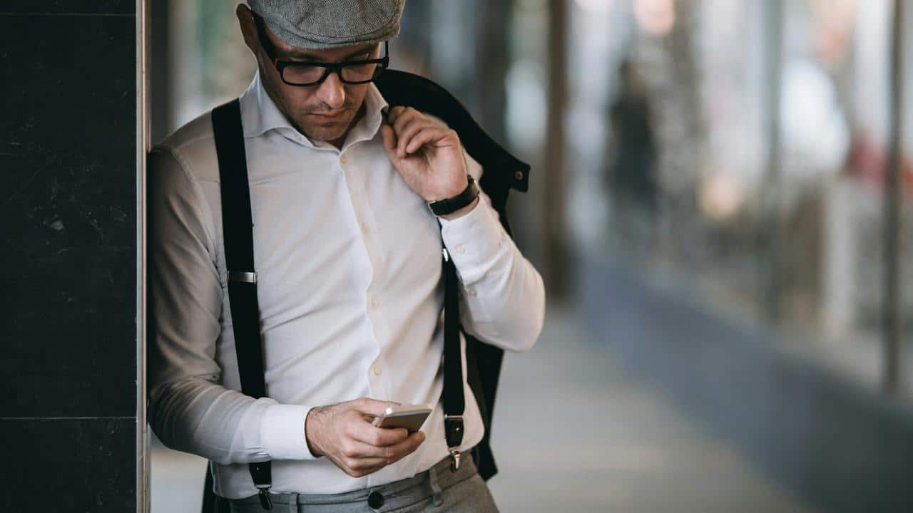 A stylish man looking at his phone while leaning against a wall.
