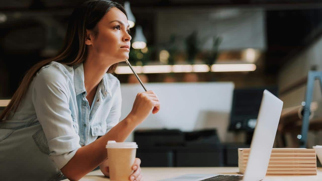 A woman holding a pencil and coffee cup while thoughtfully looking away from her laptop.