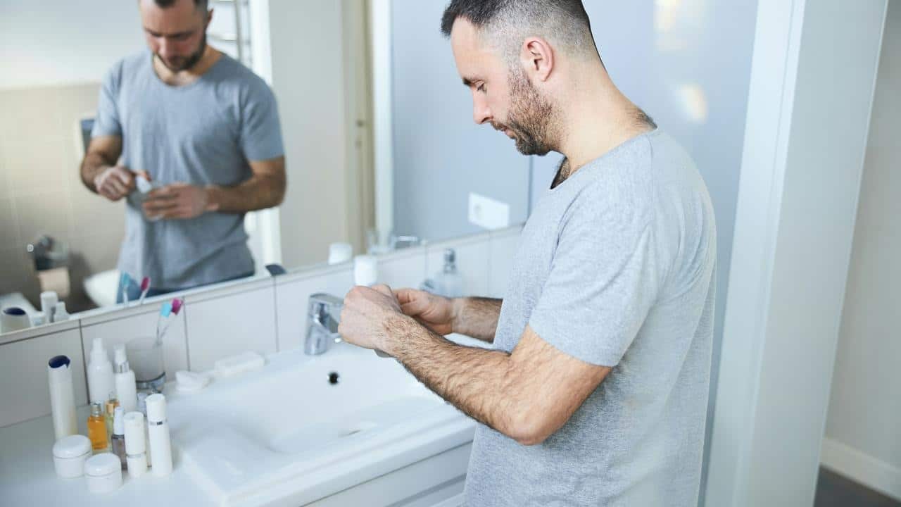 A man in a grey t-shirt using grooming products at a bathroom sink.
