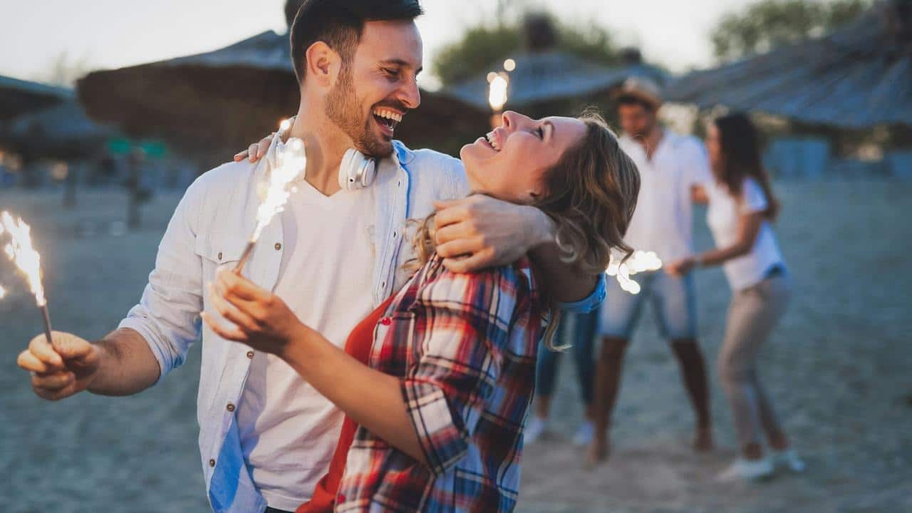 A couple laughing and holding sparklers on a beach with friends in the background.