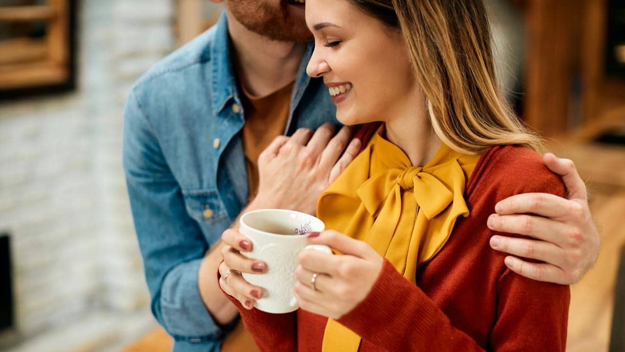 A woman holding a mug as a man hugs her from behind.