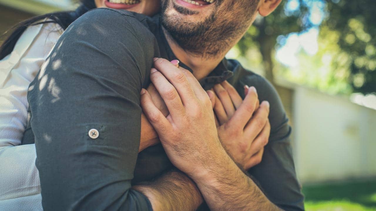 A woman hugging a man from behind outdoors.