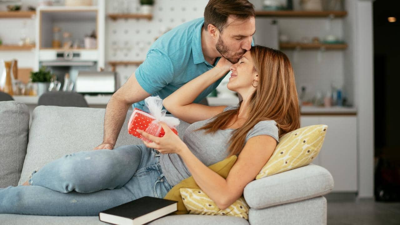 A man kissing a woman on the forehead as she holds a wrapped gift on a couch.