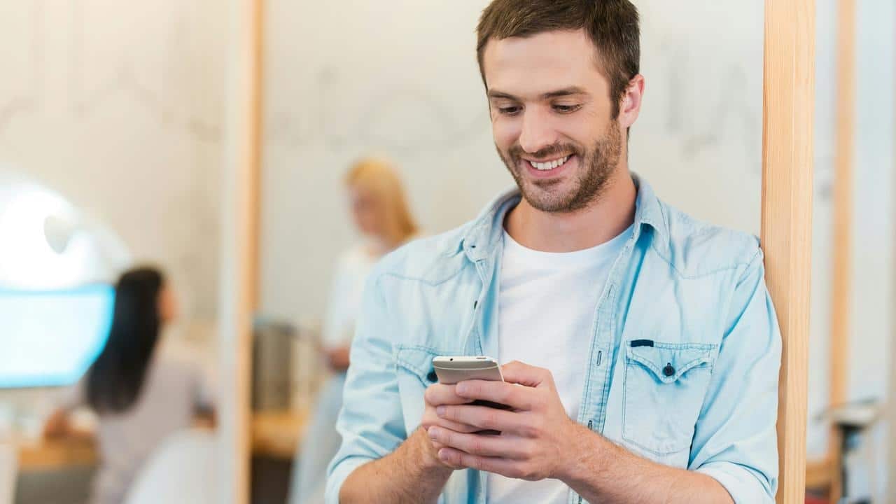 A man smiling as he checks his phone indoors.